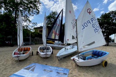 Powiększ obraz: fot. sindbadsail.com. Na zdjęciu jachty żaglowe na plaży, baner reklamowy SINDBAD sailing z informacjami o ofercie, o danych adresowych firmy położony na piasku, w tle budynek mieszkalny, drzewa