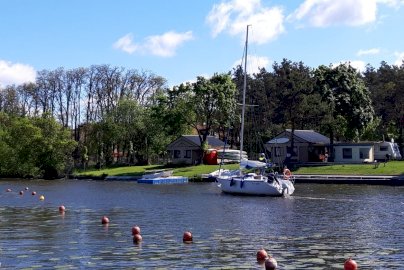 Powiększ obraz: fot. www.sailingschool.pl/. Na zdjęciu łódź pływa po wodzie. W tle domki campingowe, kamper