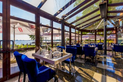 Powiększ obraz: fot. www.hotel500.com.pl. Na zdjęciu Restauracja w Zegrzu Hotel 500, zastawa na stołach, kieliszki, niebieskie fotele, złożone serwetki, przeszklone okna restauracji, na zewnątrz parasole, taras, leżaki, widok na Jezioro Zegrzyńskie