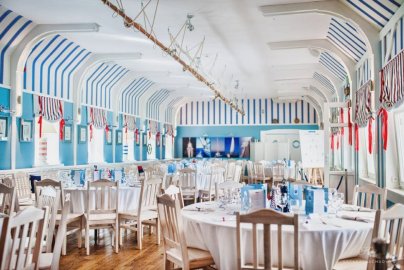 Powiększ obraz: fot. www.klubmila.pl/klub-mila-zegrzynek/restauracja. Na zdjęciu restauracja Kapitańska Klubu MIla w Zegrzynku, okrągłe stoły z krzesełkami, okna przysłonięte ozdobnymi zasłonami, karta menu na stołach