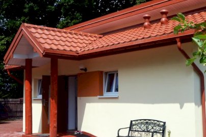 Powiększ obraz: fot. www.nocleginadzalewem.pl/index.php. Na zdjęciu widok domku z zewnątrz, otwarte drzwi wejściowe, ławka przy domu