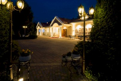 Powiększ obraz: fot. https://salamelodia.pl. Na zdjęciu Hotel Melodia Sala Bankietowa & Pensjonat. Na zewnątrz oświetlenie hotelu, kwiaty w doniczkach