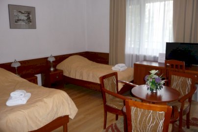 Powiększ obraz: fot. www.kss.gov.pl/dzialalnosc/dzialalnosc-informacyjno-edukacyjna/-/asset_publisher/i8pY/content/filia-kss-w-bialobrzegach?redirect=http%3A%2F%2Fwww.kss.gov.pl%2Fdzialalnosc%2Fdzialalnosc-informacyjno-edukacyjna%3Fp_p_id%3D101_INSTANCE_i8pY%26p_p_lif. Pokój hotelowy z dwoma łózkami pojedynczymi, stolikiem z krzesłami, telewizorem