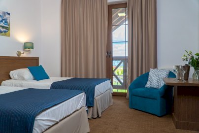 Powiększ obraz: fot. www.cichawodanieporet.pl/hotel/. Na zdjęciu pokój hotelowy z dwoma pojedynczymi łóżkami, poduszki na łóżku, niebieski fotel z poduszką przy stoliku, na stoliku kieliszki, szyjka szampana, kwiaty w wazonie, odsłonięte prawe skrzydło okienne