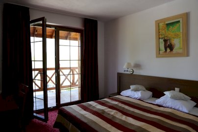 Powiększ obraz: fot. www.cichawodanieporet.pl/hotel/. Na zdjęciu pokój hotelowy z dużym łóżkiem, poduszkami do spania, obraz na ścianie, otwarte lewe drzwi balkonowe na taras, fragment stolika z krzesłem