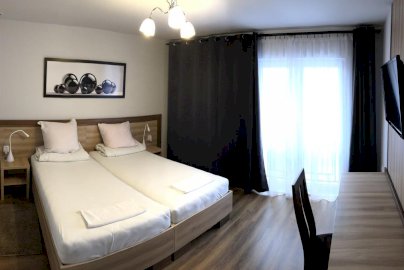 Powiększ obraz: fot. Hotel i Restauracja Prezydencka, www.facebook.com/prezydenckahr/. Na zdjęciu pokój hotelowy z złączonymi łóżkami, poduszki, pościel na łóżkach, stoliki z lampkami po obydwu stronach łóżek, po prawej stronie biurko z krzesłem oraz telewizor na ścianie