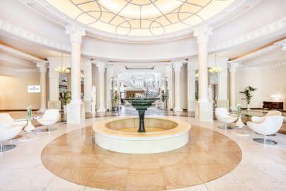 Powiększ obraz: Fot. www.windsorhotel.pl. Na zdjęciu wnętrze Hotelu Windsor, fontanna w środku, białe stoliki z fotelami, stojące rzeźby