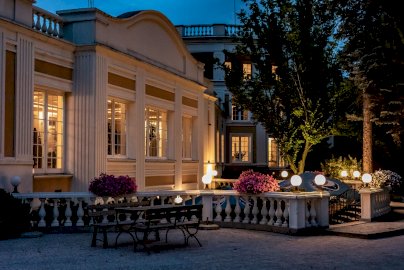 Powiększ obraz: Fot. www.windsorhotel.pl. Na zdjęciu Hotel Windsor w Jachrance wieczorową porą, lampy oświetleniowe , stolik z ławkami