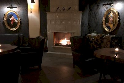 Powiększ obraz: Fot. www.hotelpantadeusz.pl. Sala z kominkiem, w kominku rozpalone drewno, stoliki z fotelami, świeczki na stolikach, obrazy w ramach na ścianie