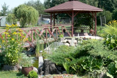 Powiększ obraz: fot. www.agroturystykajadwisin.mazowsze.pl. Na zdjęciu drewniane zadaszenie, pod nim stolik z ławkami, pomost, oczko wodne, zieleń