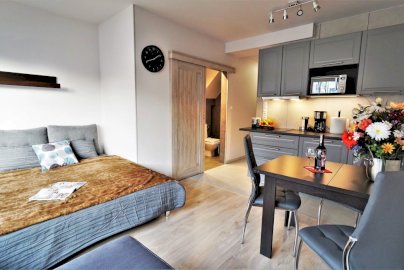 Powiększ obraz: fot. www.apartamentyserock.pl. Na zdjęciu kuchnia z wyposażeniem kuchennym: mikrofalówka, zaparzacz do kawy, stół z dwoma krzesłami, na stole bukiet kwiatów, butelka wina z kieliszkami, po lewej stronie łóżko sypialne z poduszkami