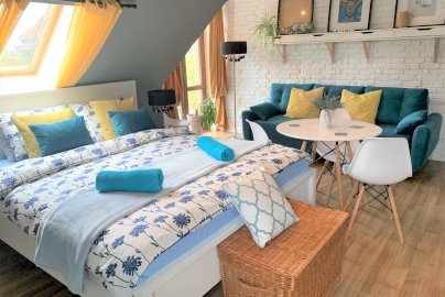 Powiększ obraz: fot. www.magnolia-rooms.pl/. Duże łóżko z poduszkami i ręcznikami, obok sofa, stolik z krzesełkami, wiklinowy schowek na ubrania