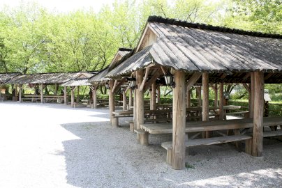 Powiększ obraz: fot. www.warszawianka.pl/pl/restauracje/miasteczko-piknikowe. Na zdjęciu miasteczko piknikowe, pod drewnianym zadaszeniem stoły z ławkami do siedzenia, w tle drzewa