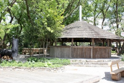 Powiększ obraz: fot. www.warszawianka.pl/pl/restauracje/miasteczko-piknikowe. Na zdjęciu altana, gdzie w środku możemy zorganizować grilla, usiąść, obok wóz z kołami, wędzarka z kominem, z tyłu drzewa