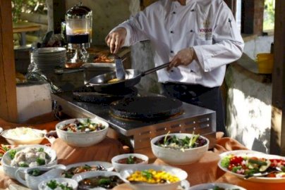 Powiększ obraz: fot. windsorhotel.pl. Na zdjęciu kucharz trzyma patelnię jedną ręką, a w drugiej trzyma nóż, z przodu przekąski, sałatki na stole obok kucharza, od góry zapalone lampy