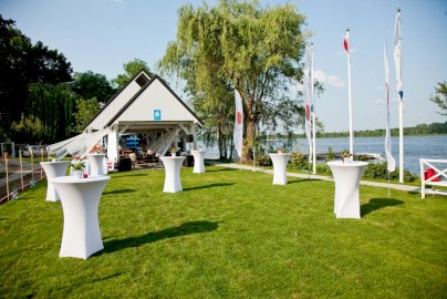 Powiększ obraz: fot. www.klubmila.pl. Na zdjęciu wysokie białe stoliki, na stolikach kwiaty, z tyłu obiekt restauracyjny, po prawej stronie rzeka Narew, pomost