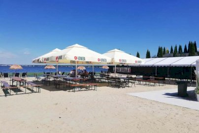 Powiększ obraz: fot. www.magnacka.pl. Na zdjęciu stoły z ławkami do siedzenia obok Jeziora Zegrzyńskiego, osoby odpoczywają siedząc na ławce pod parasolami