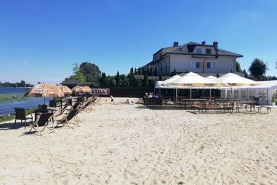 Powiększ obraz: fot. www.magnacka.pl. Na zdjęciu hotel MAGNACKA Sala gościnna, plaża na terenie hotelu, przy brzegu Jeziora Zegrzyńskiego stoliki z krzesełkami i leżakami, po prawej stronie parasole, pod nimi stoliki, ludzie