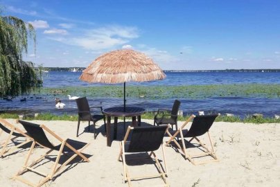 Powiększ obraz: fot. www.magnacka.pl. Na zdjęciu stolik z krzesełkami i leżakami na plaży, pływające łabędzie na tafli wody Jeziora Zegrzyńskiego
