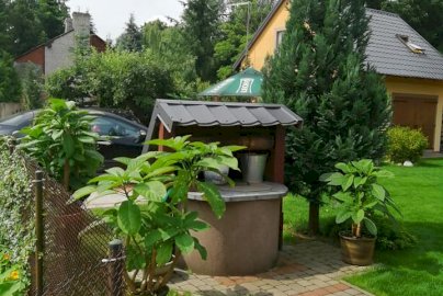 Powiększ obraz: fot. facebook Agroturystyka Pokoje u Eli, www.facebook.com/Agroturystyka-Pokoje-u-Eli-111982543878392/. Na zdjęciu studnia, czarny samochód, dom, ogród z kwiatami