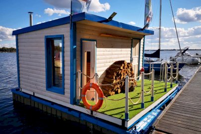 Powiększ obraz: fot. facebook Have Fun, https://www.facebook.com/havefunpl. Na zdjęciu budynek, w którym mieści się sauna, obok budynku mały skład drewna. Suna pływająca na wodzie, obok łodzie, pomost pływający