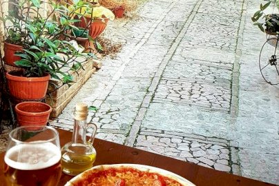 Powiększ obraz: fot. www.przystanekpizza.eu/galeria. Na  zdjęciu pizza, w kieliszku piwo, w butelce oliwa z oliwek, w tle chodnik , kwiaty w doniczkach