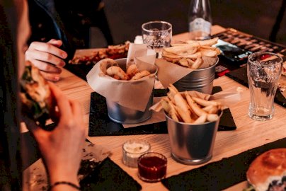 Powiększ obraz: fot. www.trustburger.pl/galeria. Na zdjęciu dziewczyna jedząca  burgera, frytki, szklanki na stole