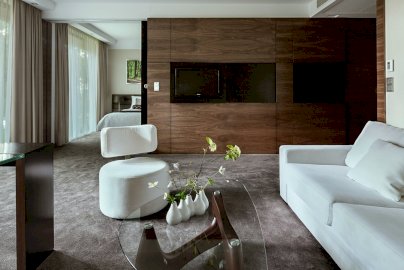 Powiększ obraz: fot. www.hotelnarvil.pl/galeria. Na zdjęciu pokój hotelowy, łóżko z poduszkami, fotel do siedzenia w pokoju, przeszklony stolik, na nim małe wazony z kwiatami, telewizor, w tle z tyłu fragment łóżka z poduszkami, obraz wiszący na ścianie