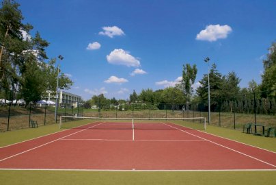 Powiększ obraz: fot. www.warszawianka.pl/pl/. Na zdjęciu kort do tenisa ziemnego, siatka, obok stolik z krzesełkami, w tle budynek, drzewa