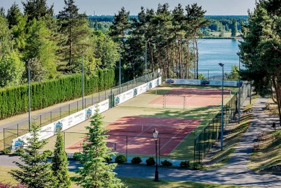 Powiększ obraz: fot. www.warszawianka.pl/pl/. Na zdjęciu boiska do gry w tenisa ziemnego, bramki do gry w piłką nożną, w tle z tyłu rzeka Narew, lasy