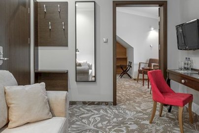 Powiększ obraz: fot. http://www.nosselia.pl/hotel/. Na zdjęciu po lewej stronie kanapa z poduszkami, z tyłu kanapy drzwi wejściowe, wieszak na ścianie, po prawej stronie stolik z czerwonym krzesełkiem, na ścianie ekran telewizora, otwarte drzwi, w tle fotel