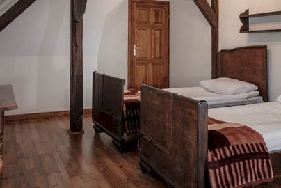 Powiększ obraz: fot. http://www.nosselia.pl/hotel/. Na zdjęciu dwa pojedyncze łóżka z poduszkami i kocami, z tyłu stolik, na stoliku lampka, drewniane krzesełko obok stolika, nieduża półka nad łóżkami