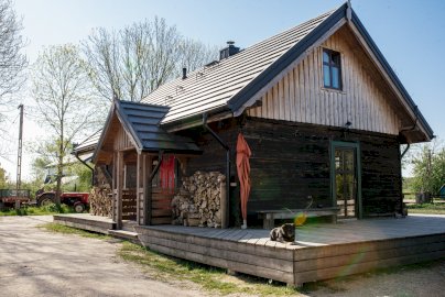 Powiększ obraz: fot. Chatka Antka, https://siedliskoleluja.pl/nocleg/#!/chatka-antka. Na zdjęciu drewniana chata, obszerny taras przed domem, pies leży na tarasie, z tyłu mężczyzna kieruje traktorem