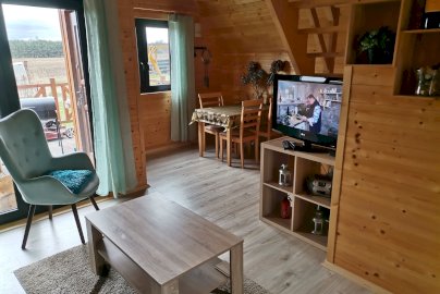 Powiększ obraz: fot. www.dzikizakatekpiskornia.pl. Na zdjęciu domek nad Narwią w Dzierżeninie. Na zdjęciu wnętrze pokoju w domku w Dzierżeninie, na szafce telewizor, z tyłu stolik z krzesełkami, za oknem balkonowym grill, widok na pole i las