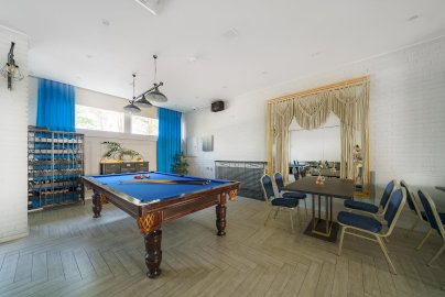 Powiększ obraz: fot. facebook Marina Diana, https://www.facebook.com/HotelMarinaDiana?locale=pl_PL. Na zdjęciu stół do bilarda z kijami i bilami na stole, w suficie wisi lampa z trzema kloszami, przy ścianie półki, szafka z szufladami, na ścianie przy suficie głośnik, po prawej stronie stoliki z krzesełkami, mężczyzna siedzący z tyłu za zasłoną
