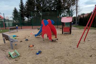 Powiększ obraz: fot. Krzysztof Kozań. Na zdjęciu piaskownica, zabawki dla dzieci leżą na piasku w piaskownicy, krótka zjeżdżalnia dla dzieci, ławki, huśtawka, z tyłu boisko do gry w piłkę koszykową, stół do tenisa stołowego