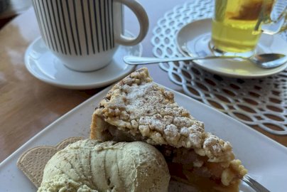 Powiększ obraz: fot. facebook Cafe Verona Legionowo, https://www.facebook.com/CafeVerona/?locale=pl_PL. Na zdjęciu na talerzyku szarlotka, gałki lodów, ciastka, kubek, wysoka szklanka z jasnym naparem z herbaty, długa łyżeczka do mieszania na talerzyku, z tyłu dynia, kwiaty