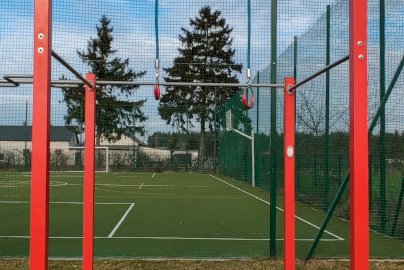 Powiększ obraz: fot. Krzysztof Kozań. Na zdjęciu drążek do podciągania się z kółkami gimnastycznymi do ćwiczeń, z tyłu drążek, fragment boiska do piłki nożnej oraz koszykowej wykonany z nawierzchni syntetycznej