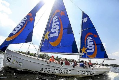 Powiększ obraz: fot. Ośrodek Sportów Wodnych PORT AZS ZEGRZE. Na zdjęciu żaglówka z granatowymi żaglami płynie po jeziorze z grupą ludzi na pokładzie