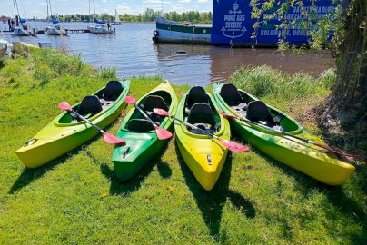 Powiększ obraz: fot. Ośrodek Sportów Wodnych PORT AZS ZEGRZE. Na zdjęciu na trawie widać cztery kajaki dwuosobowe z wiosłami. Kajaki barwy jasnozielonej i zielonej. Z tylu jezioro oraz dużej wielkości baner z napisem PORT AZS ZEGRZE. Po lewej stronie od baneru zacumowane do pomostu łodzie bez podniesionych żagli
