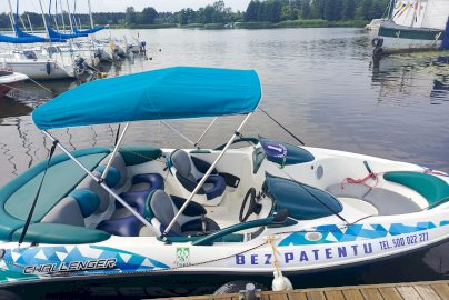 Powiększ obraz: fot. Ośrodek Sportów Wodnych PORT AZS ZEGRZE. Łódź motorowodna bez patentu stojąca na wodzie jeziora. Z tyłu po lewej stronie za łodzią motorowodną widać ustawione w rzędzie łodzie bez podniesionych żagli. W oddali rosnące drzewa