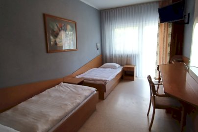 Powiększ obraz: fot. www.geovita.pl/jadwisin/. Na zdjęciu widać pokój dwuosobowy z dwoma pojedynczymi łóżkami. Po prawej stronie znajduje się stolik z krzesłem, w rogu wisi telewizor, a po lewej stronie na ścianie — obraz
