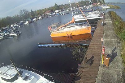 Powiększ obraz: fot. TKKF Wodnik. Widok na przystań z pomostem i zacumowanymi jachtami, na pierwszym planie pomarańczowy żaglowiec, a w tle liczne łodzie motorowe i żaglówki stojące w spokojnej wodzie