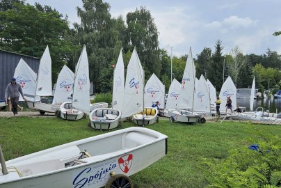 Powiększ obraz: fot. Klub Sportowy "Spójnia-Warszawa". Na trawiastym brzegu jeziora ustawionych jest wiele żaglówek Optimist należących do klubu sportowego Spójnia - Warszawa
