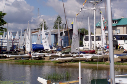 Powiększ obraz: fot. Ośrodek MOS-2, filia w Zegrzu Południowym. Na zdjęciu teren ośrodka żeglarskiego z licznymi małymi jachtami ustawionymi na brzegu i zabudowaniami portowymi w tle